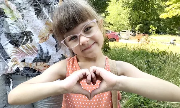 Young girl makes a heart shape with her hands while standing outdoors.